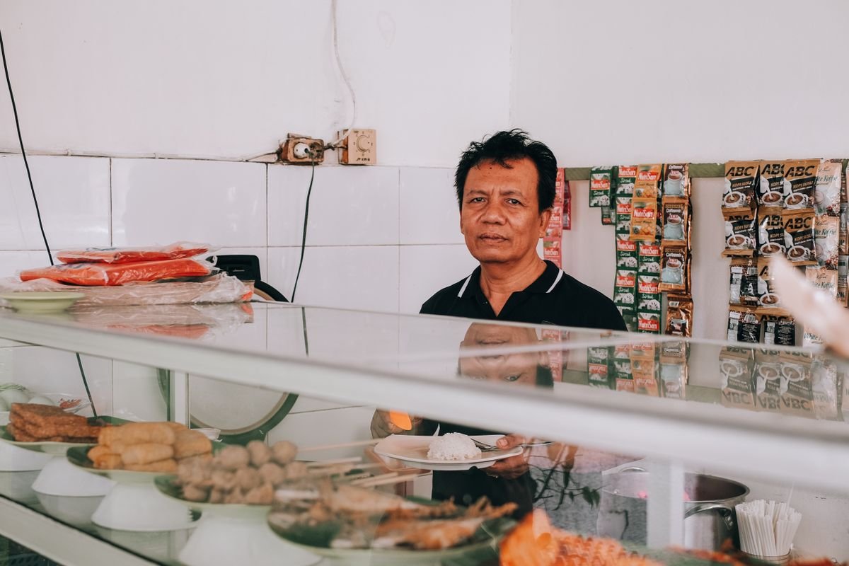 Pemilik warung makan berdiri di balik etalase kaca berisi lauk-pauk dagangannya