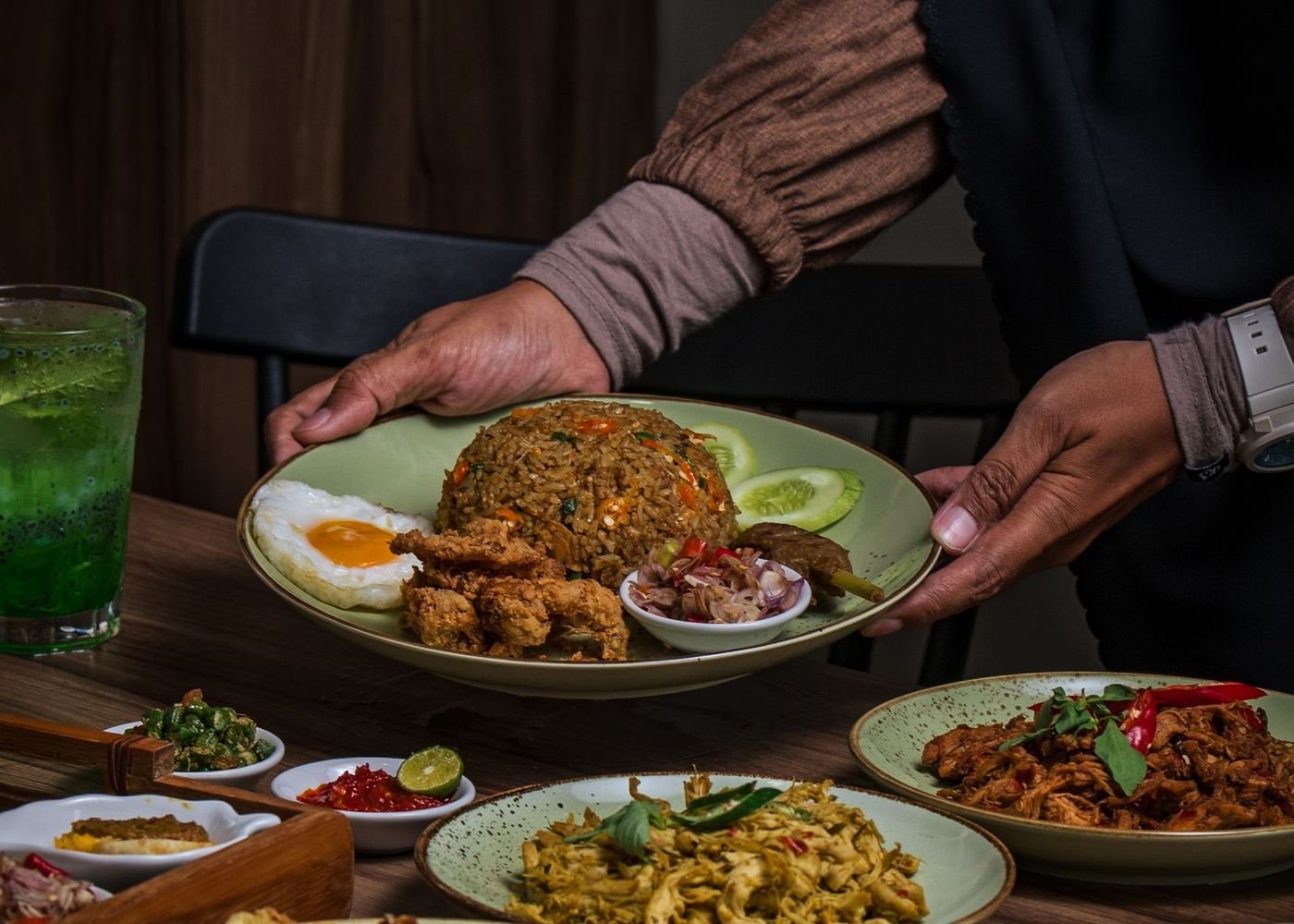 Tangan menyajikan nasi goreng dan aneka lauk di meja kayu Dapur Bu Sri
